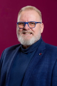 Portretfoto van Jan Posdijk. Blauw jasje, blauwe koltrui en bordeaux rode achtergrond. Fotograaf: Jaap Lotstra